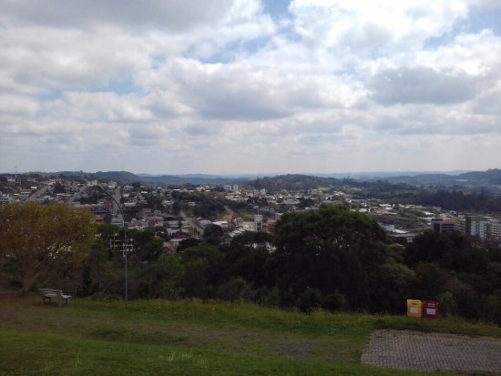 Vista panorâmica da cidade de São Marcos (RS) em um dia parcialmente nublado, com área verde em primeiro plano, árvores e bancos de praça, prédios residenciais e comerciais ao fundo, além de ruas e morros que se estendem até o horizonte.