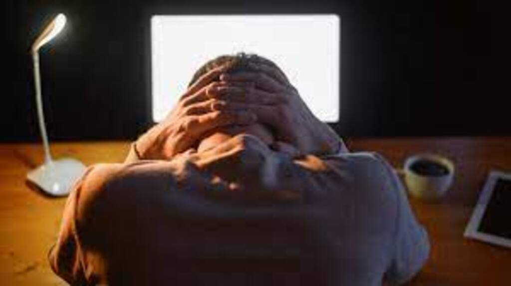 Pessoa sentada em frente a um computador, apoiando a cabeça nas mãos em sinal de cansaço ou estresse; ambiente com iluminação baixa, abajur aceso e uma xícara de café sobre a mesa.