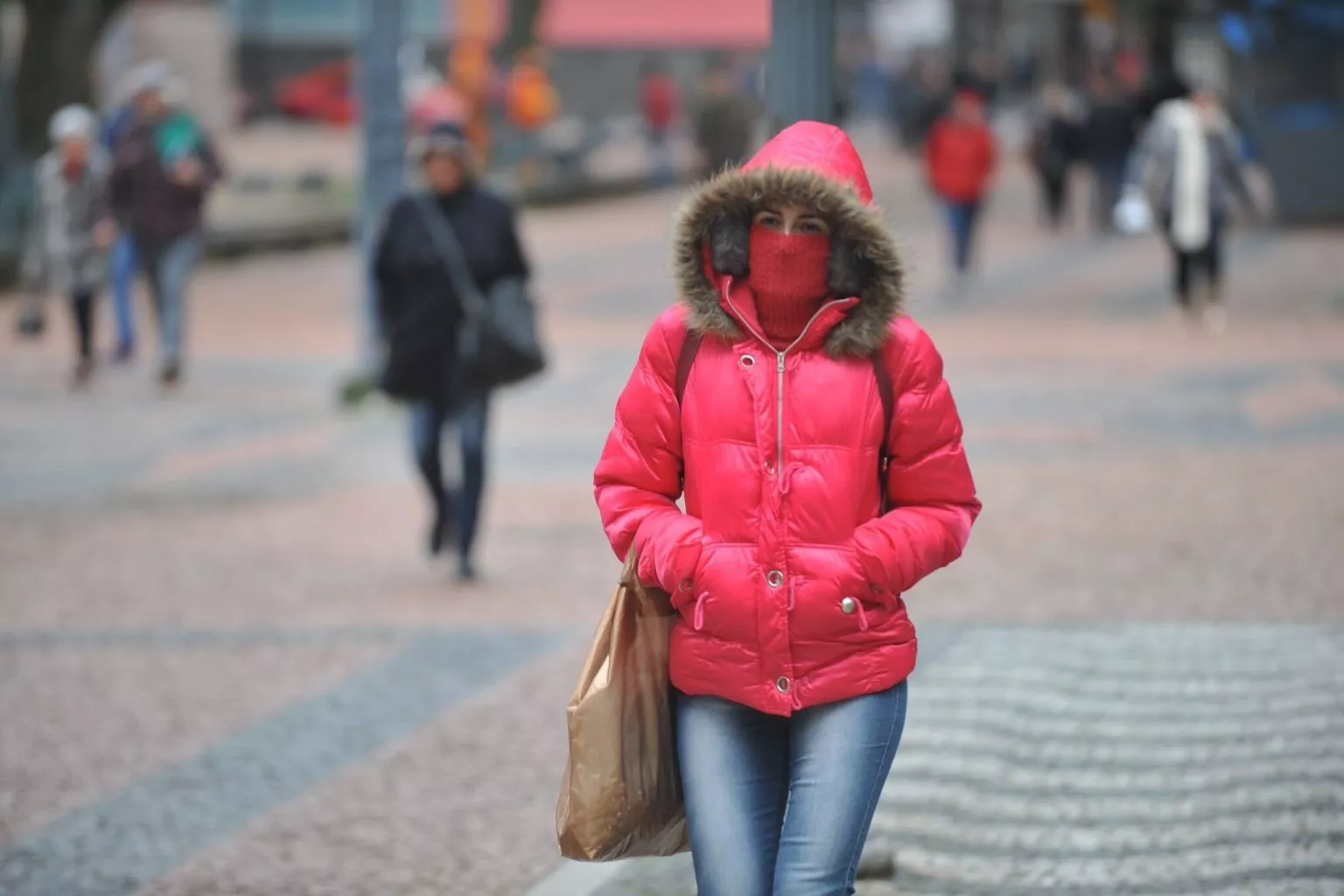 Pessoa caminha em rua durante dia frio, vestindo casaco de inverno vermelho com capuz de pele e cachecol cobrindo parte do rosto. Outras pessoas aparecem ao fundo também agasalhadas.