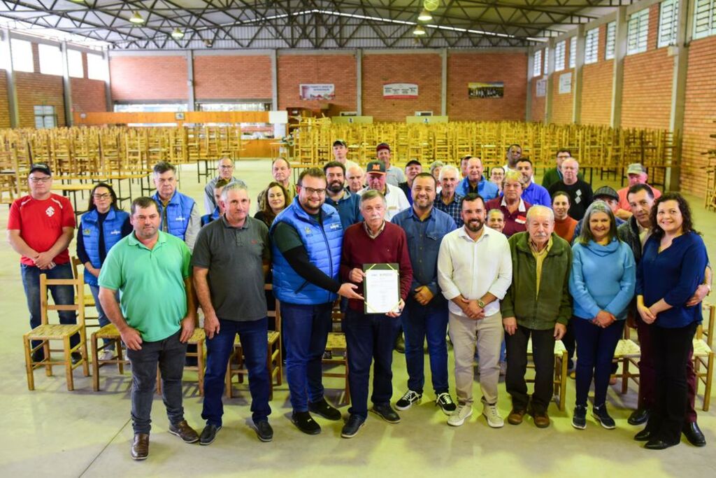 Foto mostra um grupo de moradores, autoridades e representantes do Samae reunidos em um salão comunitário em Criúva. Ao centro, um morador segura o documento que autoriza a perfuração do poço artesiano. Ao fundo, diversas cadeiras estão empilhadas, e o espaço é iluminado por luz natural e artificial. | Foto: Laura Piola