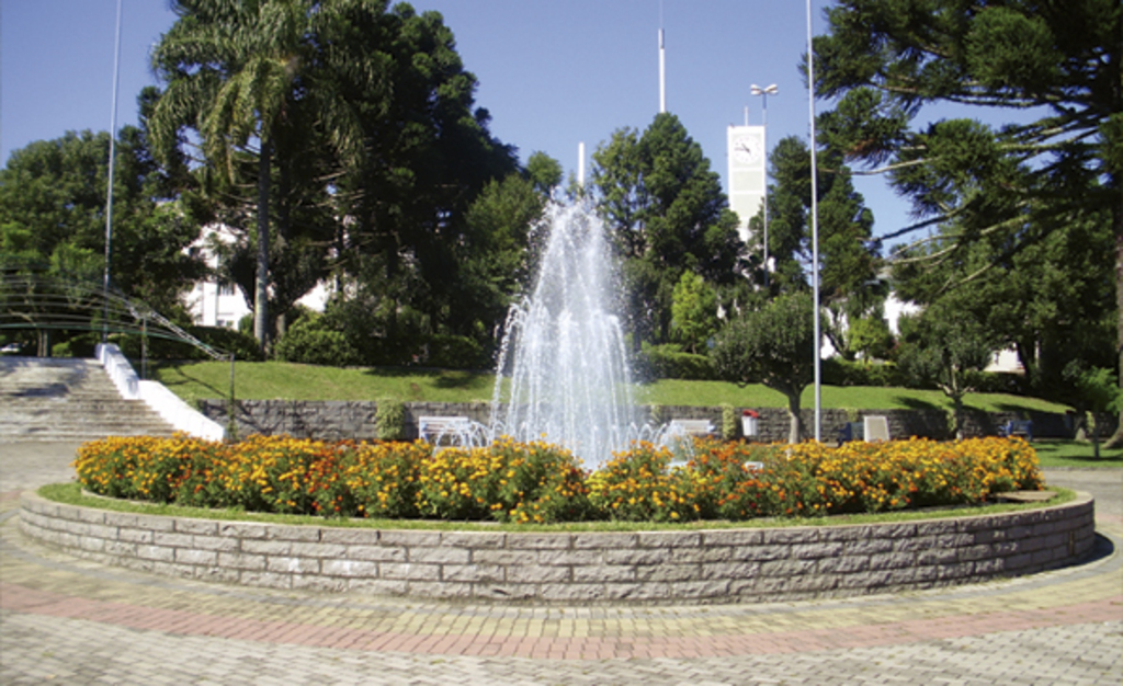 Praça central de São Marcos, RS, com fonte de água em funcionamento cercada por um canteiro de flores coloridas. Ao fundo, árvores de grande porte e o prédio da prefeitura com torre do relógio visível. Céu limpo e ensolarado.