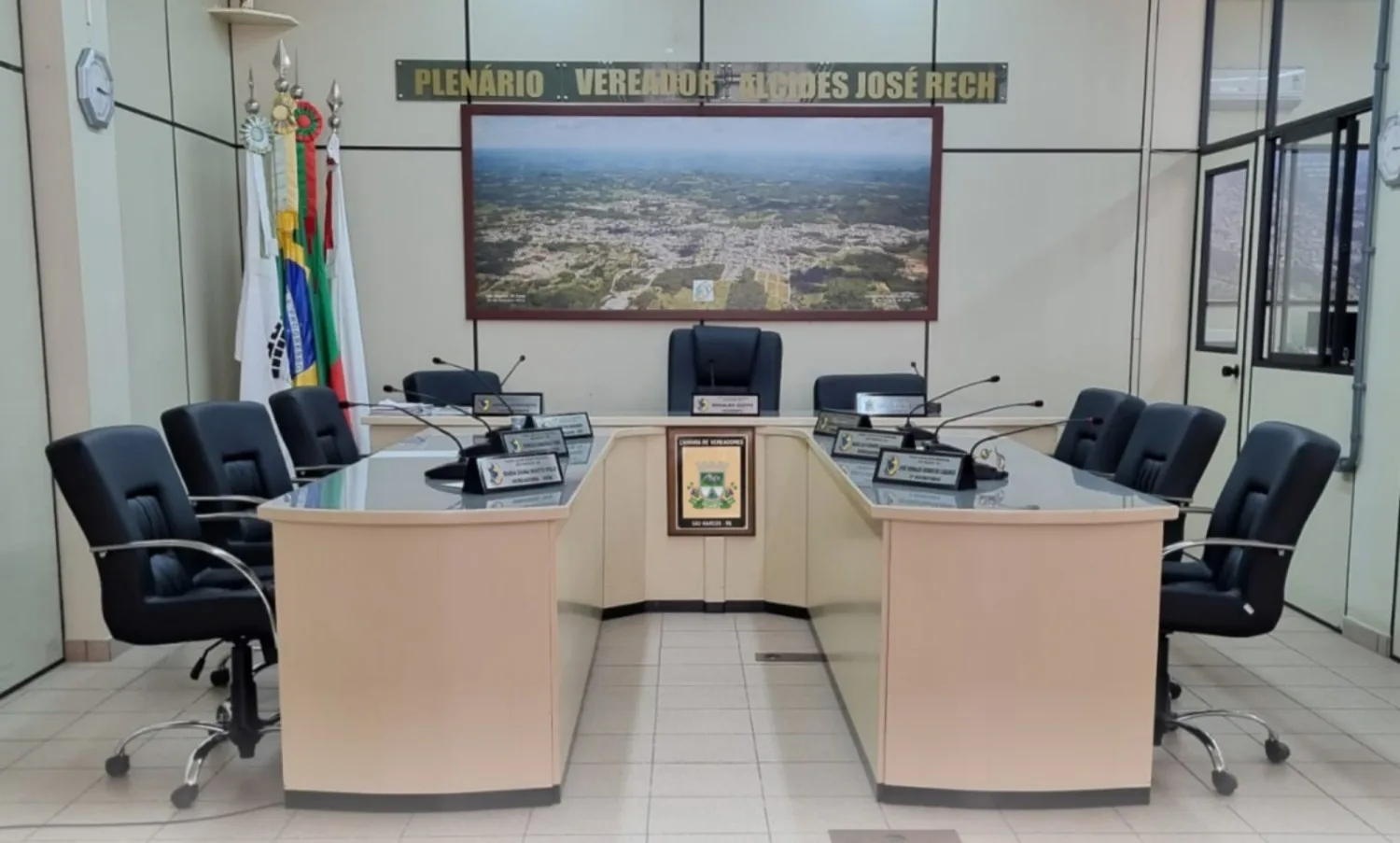 Plenário da Câmara de Vereadores de São Marcos, com cadeiras e microfones dispostos em formato semicircular, bandeiras ao fundo e imagem aérea da cidade na parede.