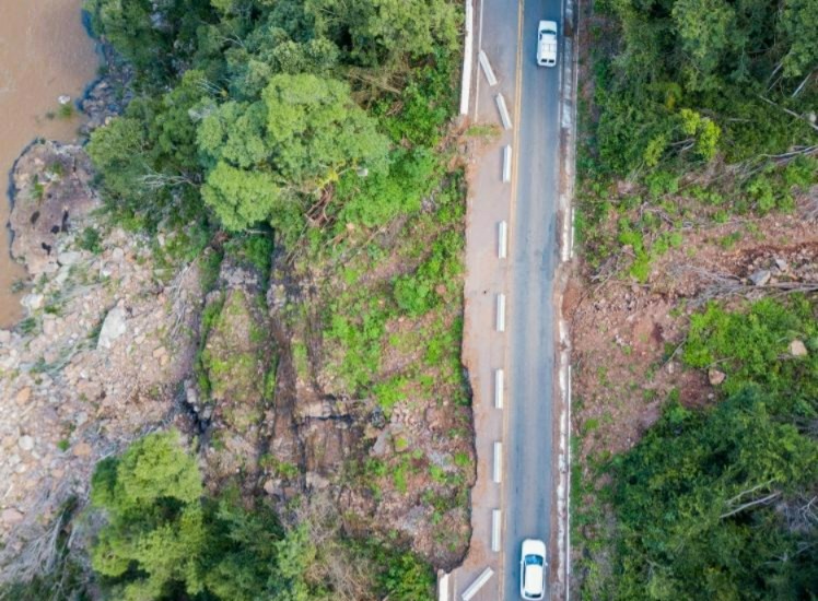 Imagem aérea do km 96 da BR-116, em São Marcos (RS), mostra trecho onde metade da pista foi destruída por uma penetração de terra. À esquerda, encosta íngreme com superfícies e rochas expostas próximas ao leito do Rio das Antas. À direita, a pista remanescente segue estreita e com trânsito controlado por semáforo, com dois veículos circulando. A imagem evidencia a gravidade da penetração e os riscos à infraestrutura da rodovia. Créditos: viagensecaminhos