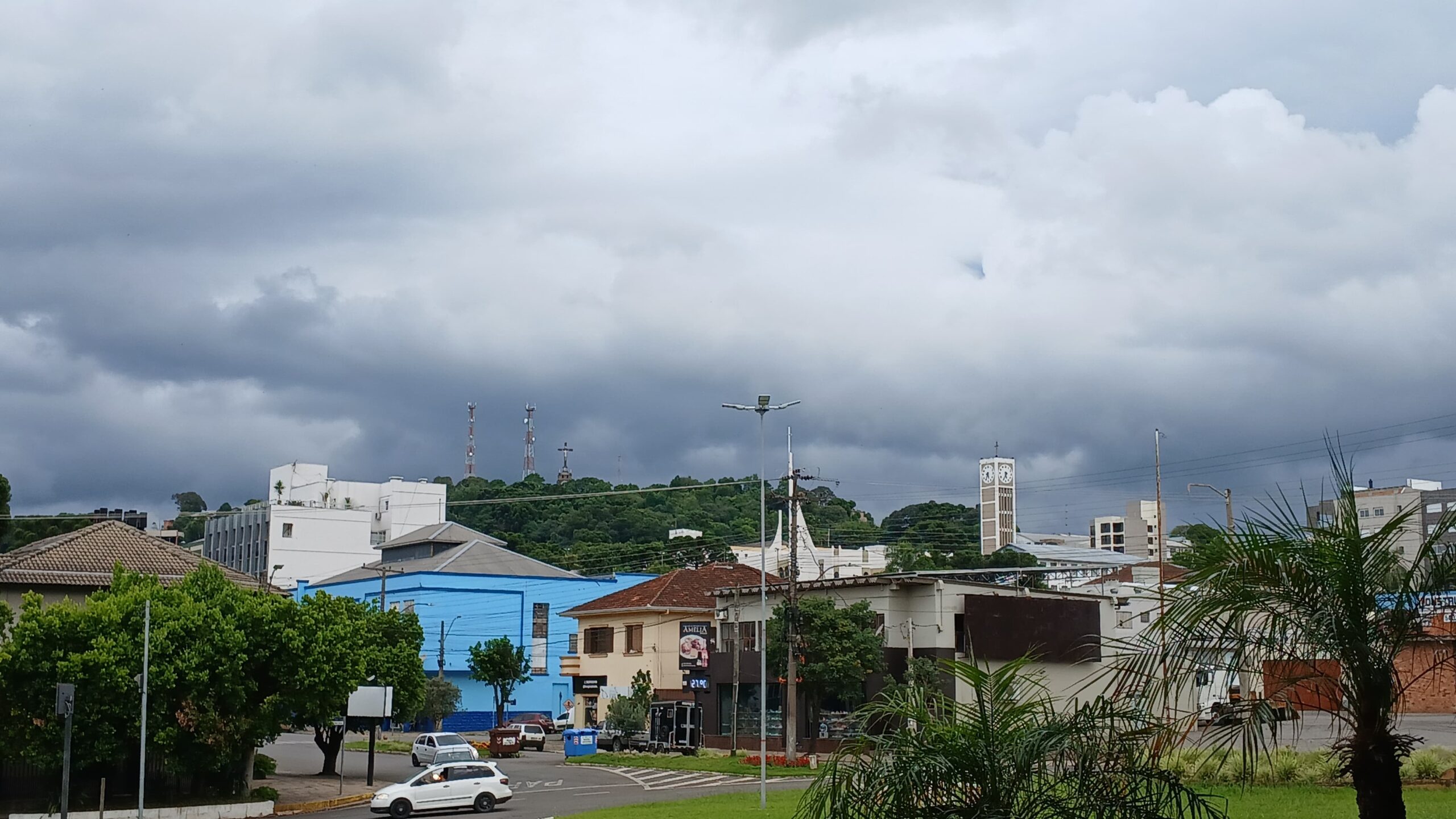Vista urbana de São Marcos (RS), com céu carregado de nuvens cinzas e árvores e prédios ao fundo, com destaque para a Torre da Matriz e o Monte Calvário. Créditos: São Marcos Online.